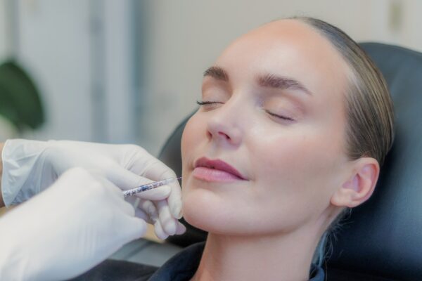 Behandler giver botox langs kæbelinjen med en tynd insulinsprøjte, kvinden ligger med lukkede øjne i den sorte behandlingsstol