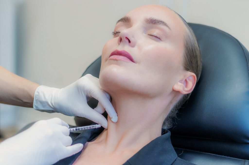 Behandler giver botox i halsen med en tynd insulinsprøjte, kvinden læner hovedet tilbage i behandlingsstolen